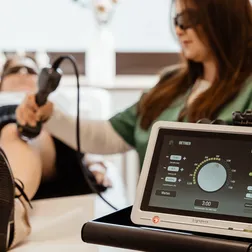 Eine Sprechstundenhelferin mit Schutzbrille setzt bei einer Patientin das Lasergerät am Knie an