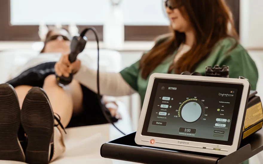 Eine Sprechstundenhelferin mit Schutzbrille setzt bei einer Patientin das Lasergerät am Knie an