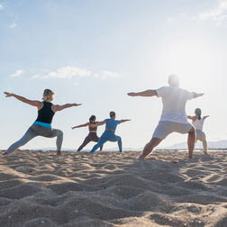 Gegenlichtaufnahme einer sechsköpfigen Gruppe in Sportkleidung beim Yoga übt Kriegerpose am Strand bei Sonnenaufgang