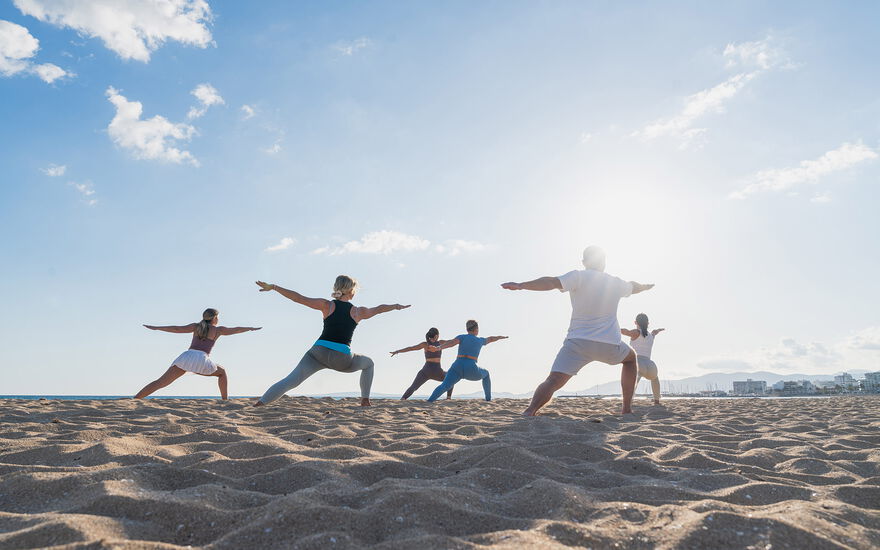 Gegenlichtaufnahme einer sechsköpfigen Gruppe in Sportkleidung beim Yoga übt Kriegerpose am Strand bei Sonnenaufgang