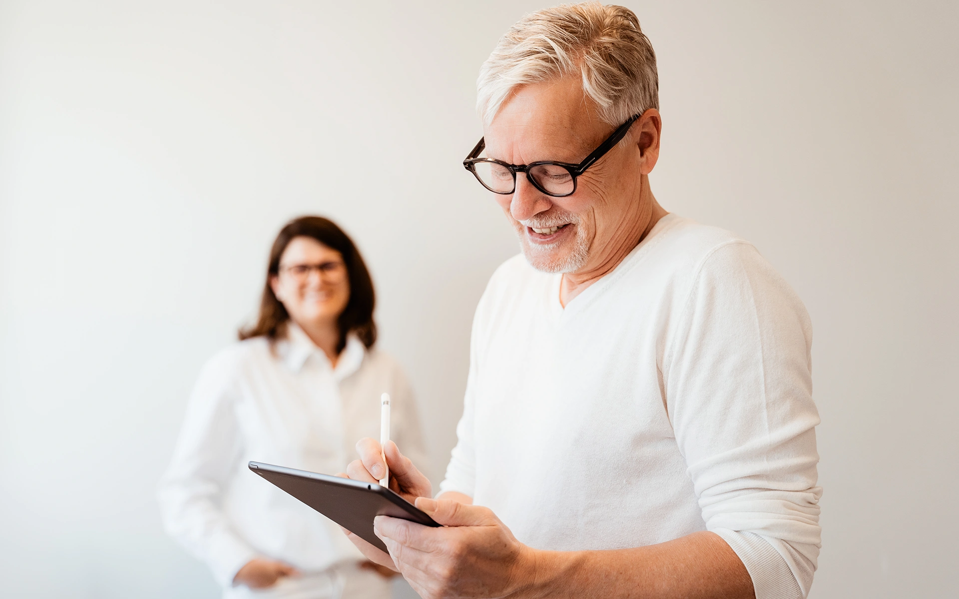 Dr. Burkhard Schubert unscharf mit iPad und Dr. Kathrin Hanswille im Hintergrund mit weißer Kleidung in der Praxis